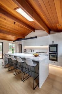 Modern kitchen with a white marble island and light gray cabinets beneath a wood-paneled vaulted ceiling; bar stools line the island