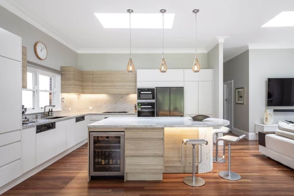 Bright modern kitchen with white cabinets, wood accents, marble countertops, and three pendant lights over a curved island.