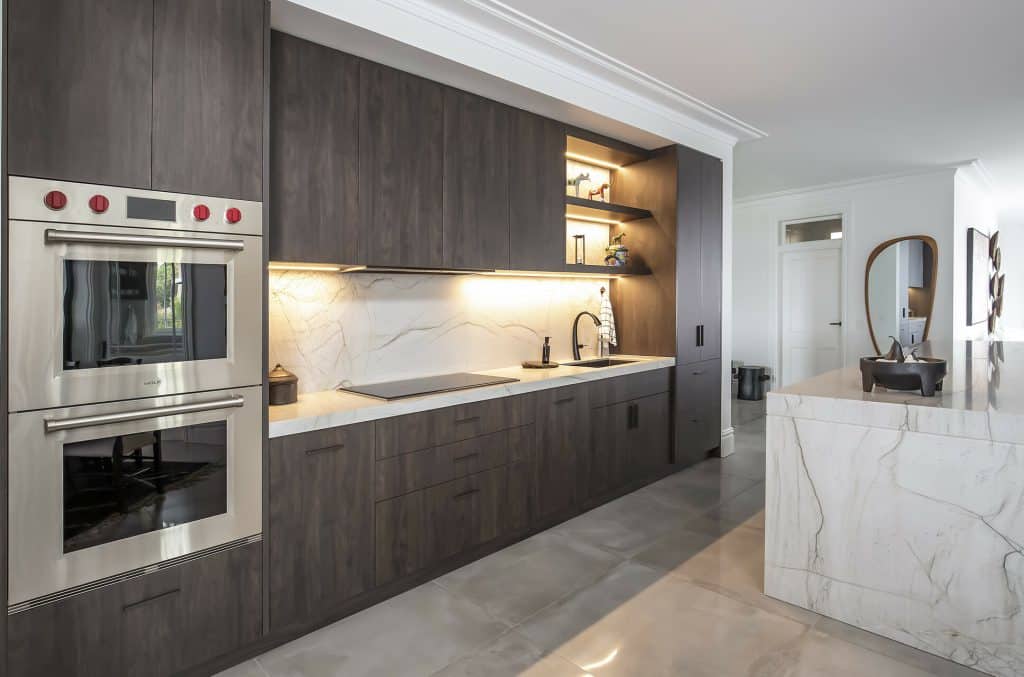 Modern kitchen with dark wood cabinets, stainless ovens, and marble countertops; open shelf lighting above the sink area.