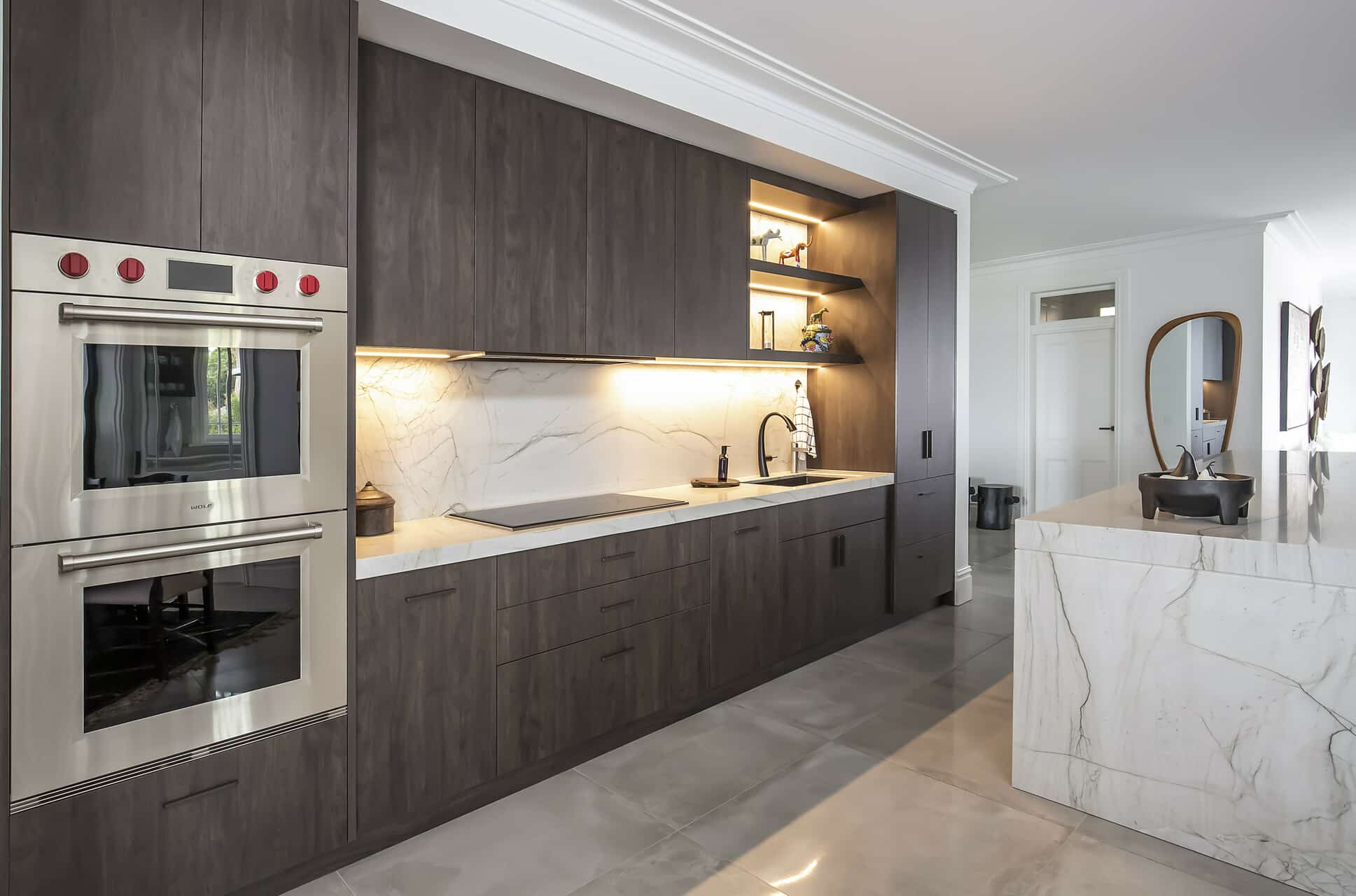 Modern kitchen with dark wood cabinets, stainless ovens, and marble countertops; open shelf lighting above the sink area.