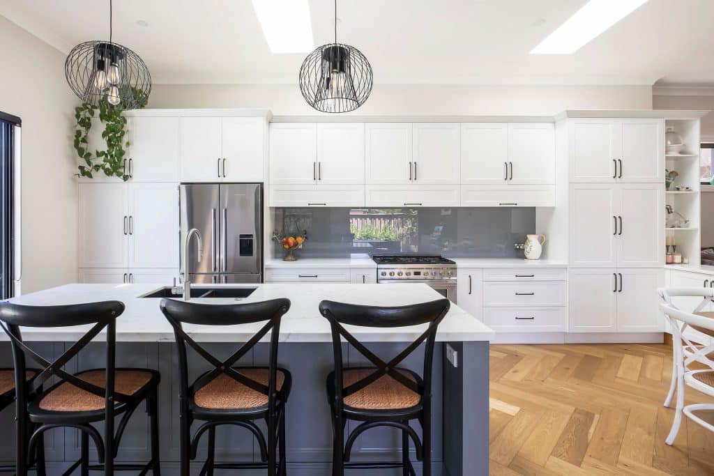 Bright white kitchen with an island, black cross-back stools, stainless steel refrigerator, and white cabinets.