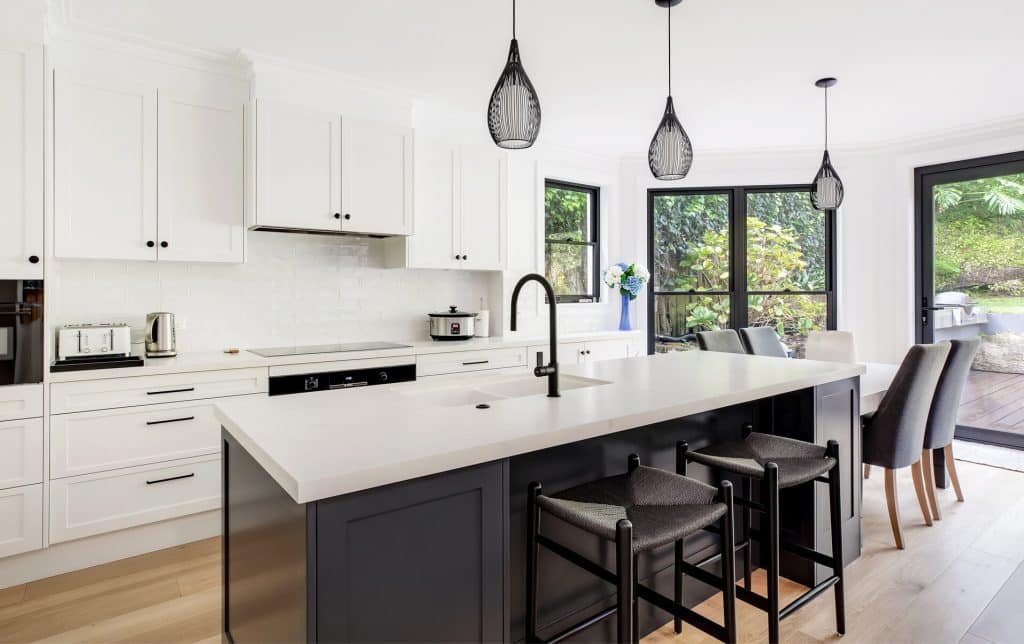 Bright white kitchen with an island, black pendant lights, and large windows showing greenery outside.
