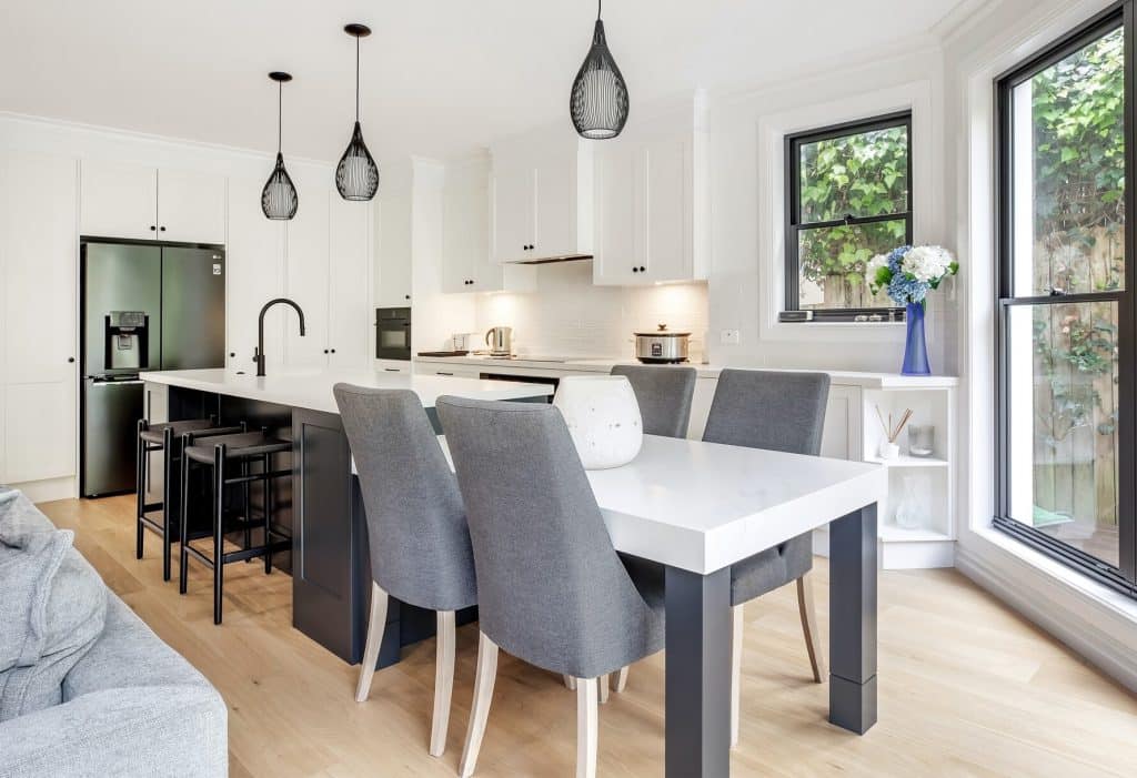 Bright modern kitchen with white cabinets and a large white island, gray dining chairs, and black pendant lights above the island.