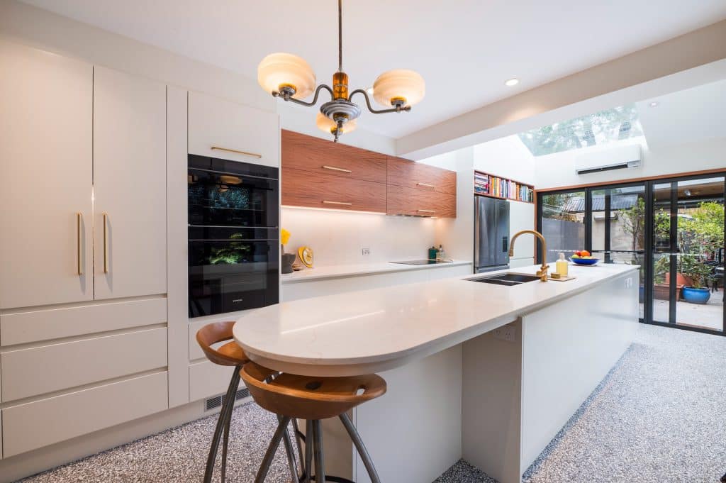 Modern kitchen with white cabinets, a marble island, brass hardware, and a wooden-accent wall above the counter. Sliding glass doors open to an outdoor patio.