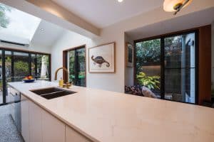 Bright modern kitchen with a white marble island, brass faucet, fruit bowl, and large glass doors opening to greenery outside.